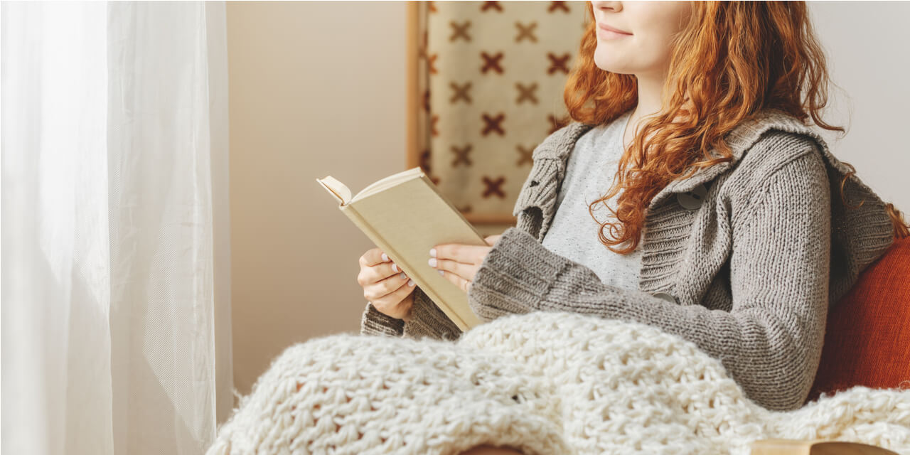 Girl with red hair sitting in a chair while reading a book, covered with a blanket