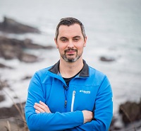 Karl Henry wearing blue top, standing with arms folded in a beach setting