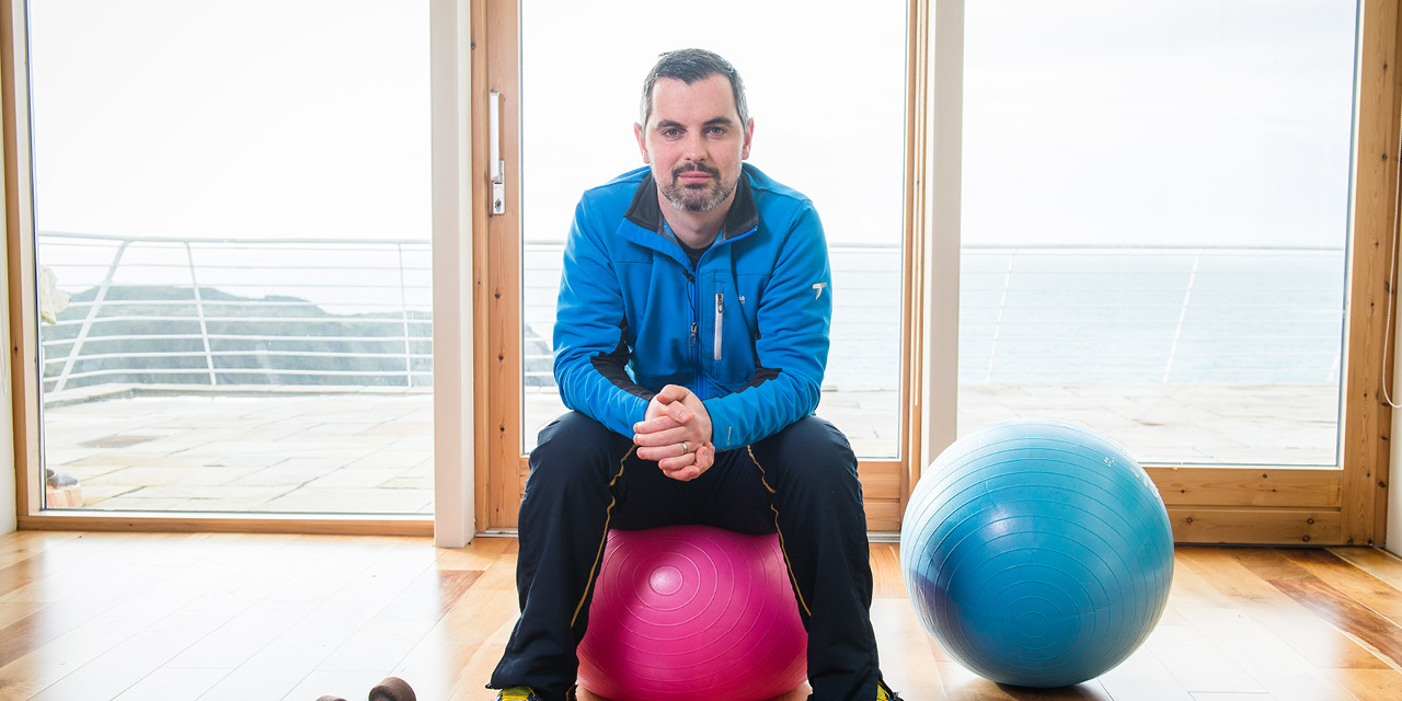 Karl Henry in a blue top, sitting on a pink exercise ball